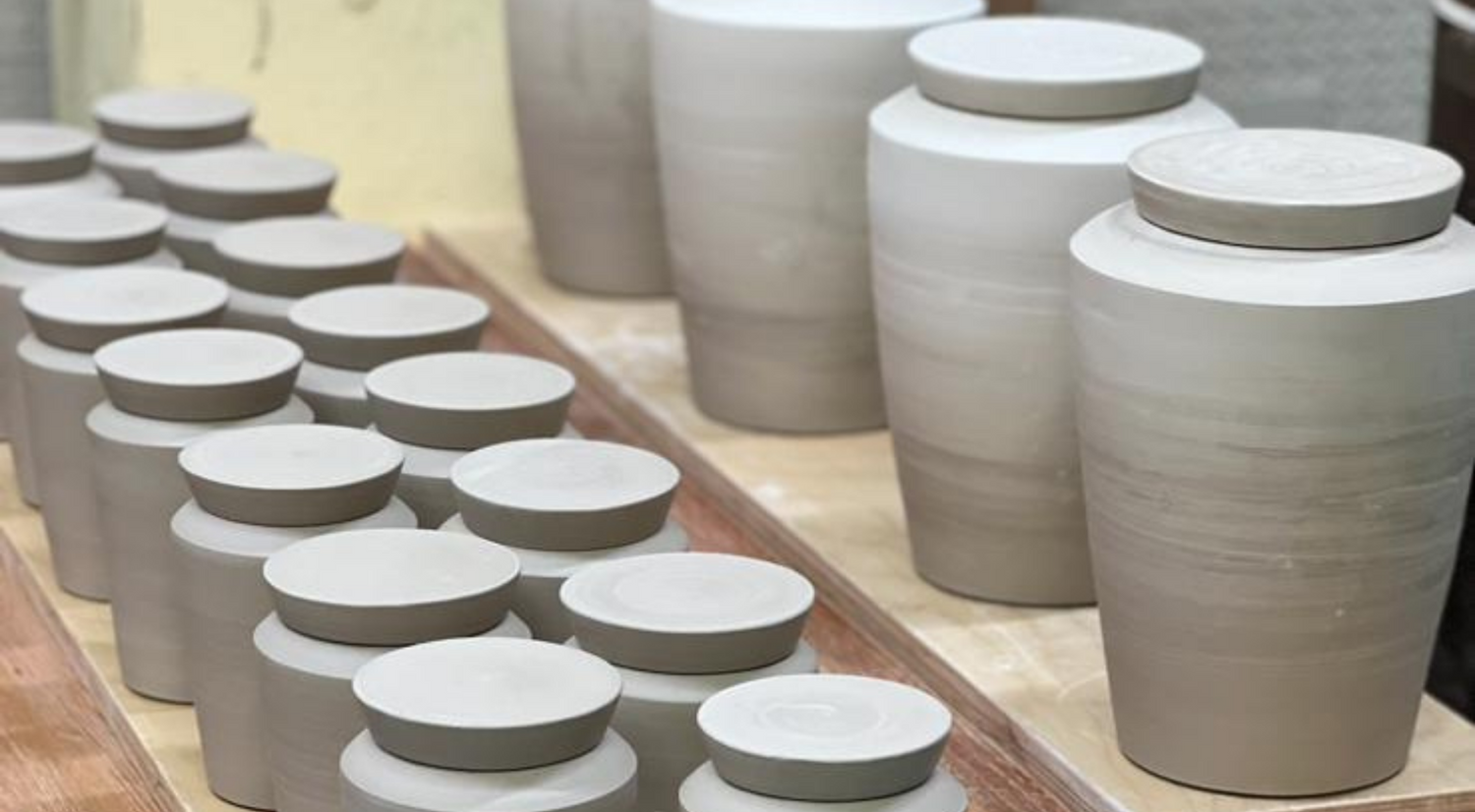 Rows of unfinished clay ceramic cremation urns drying on wooden boards in a pottery studio.