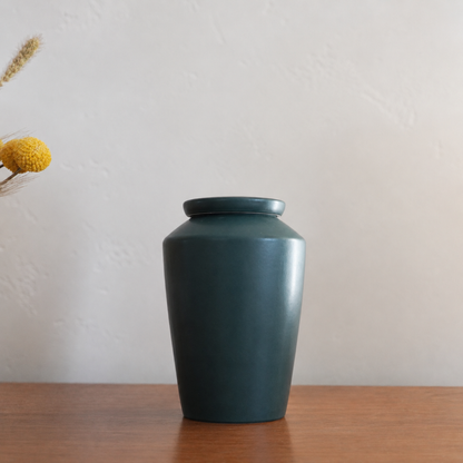 Dark green keepsake ceramic urn on a wooden shelf beside dried flowers, a discreet home memorial.