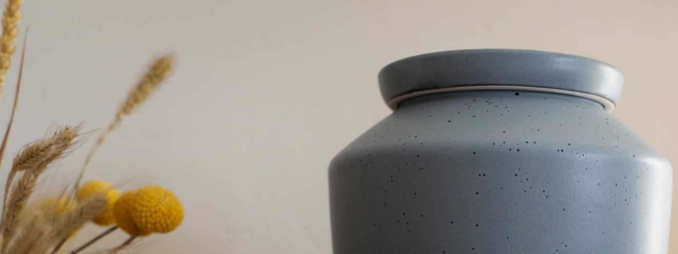 Close-up of a modern gray speckled ceramic cremation urn with yellow billy balls and dried wheat in a minimalist home setting.