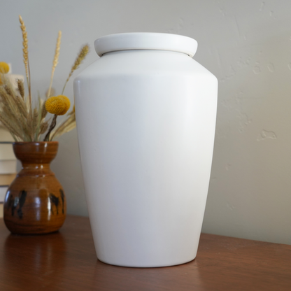 Modern matte white ceramic urn styled as an art piece on a bookshelf, a discreet and beautiful home memorial.